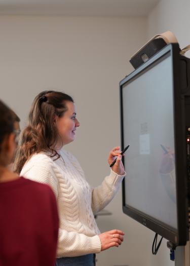 Eine junge Frau notiert etwas auf einem Smartboard, neben ihr sind junge Menschen von hinten zu erkennen